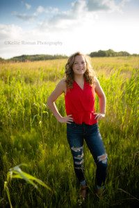 milwaukee senior pics a teenage girl from oak creek in milwaukee is standing in a field of tall green grass. she has on dark jeans with holes, and a red top. she has her hands on her hips and the sun is setting behind her .