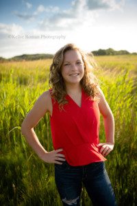 milwaukee senior pics a teenage girl from oak creek in milwaukee is standing in a field of tall green grass. she has on dark jeans with holes, and a red top. she has her hands on her hips and the sun is setting behind her .