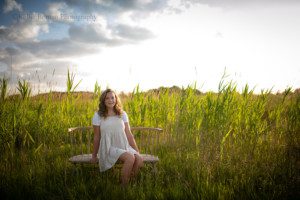 milwaukee senior pics a teenage girl from oak creek is sitting on a wood bench in a field of very tall green grass. the sun is setting behind her, and the sky is blue with a few puffy clouds. she is wearing a beige dress and a white shirt under it