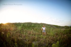 milwaukee senior pics a senior girl from oak creek Wisconsin is standing in front of a huge hill with tall grass. she's wearing a white shirt and a black and white skirt. she's got her hands on her hips and the sun is setting off to the side