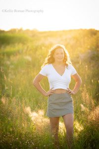 milwaukee senior pics a young teenage girl is outdoors in a Milwaukee County park. the sunset is shining from behind her and there is lots of golden sunflair on her. she's wearing a white shirt showing her stomach, with a black and white skirt and has her hands on her hips
