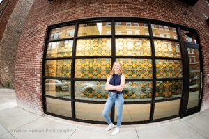 senior locations in milwaukee a high school girl from oak creek is in downtown Milwaukee in the third ward in front of a large class window with a few of a mosaic painting on the inside of the building. she's wearing a navy tank top and jeans.