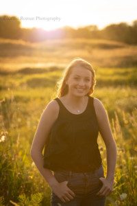 senior locations in Milwaukee a senior girl from oak creek is surrounded by sunlight and tall grass she has a black shirt on with jeans and has her thumbs in her pockets the sun in shining behind her