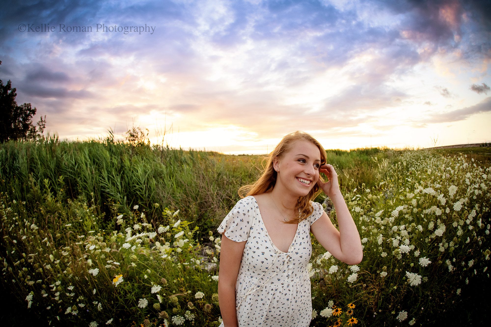 senior locations in Milwaukee a teenage girl in a Milwaukee County park she has blonde hair and has her hand running through it. she's looking off to the side smiling with tall grass surrounding her at sunset. the sky has beautiful purple and pink clouds