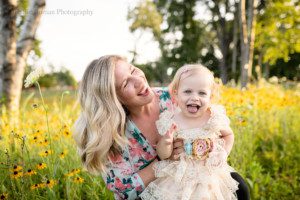 milwaukee childrens photographer. A two year old wearing a cream lace dress is sitting on her moms lap and looking while smiling at the camera. her mom has her eyes closed and is also laughing. They have a lot of yellow black eyed Susan flowers behind them.