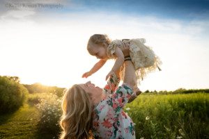 milwaukee childrens photographer. A mother is holding her two year old daughter up in the air in a field of tall grass and flowers during sunset. The mom has blonde hair and the sun is shining through it. She is smiling up at her daughter, and her daughter is reading down for her mom.