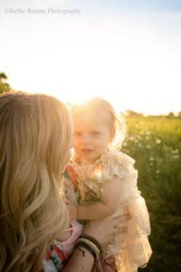 milwaukee childrens photographer. a two year old girl is being held by her mom while outside during the sunset. The girl is looking at the camera with big blue eyes, while her mom is looking at her daughter. The little girl has a beige romper on and blonde curly hair.