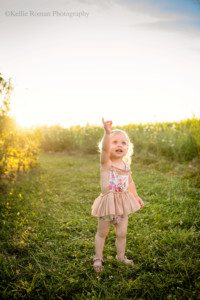 milwaukee childrens photographer. a two year old girl is standing outside with tall grass and bushes behind her. the sun is setting and it's glowing gold behind her lighting up her blonde curly hair. She has big blue eyes and is wearing a beige and pink floral romper. she's pointing and looking up at the sky.