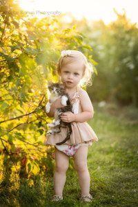 milwaukee childrens photographer. a two year old girl is standing outside with tall grass and bushes behind her. the sun is setting and it's glowing gold behind her lighting up her blonde curly hair. She has big blue eyes and is wearing a beige and pink floral romper while holding a white and grey kitten.