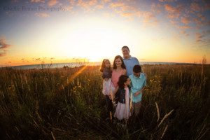 milwaukee sunrise photography. a family of five is in a field of tall grass in Milwaukee County. the sun is rising behind them and everything is glowing gold with blue and purple in the sky. two kids are looking at each other while mom dad and little sister are looking at the camera.