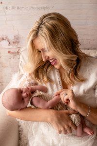 sweet and simple newborn. a new mother is holding her infant son in a milwaukee photo studio. she is holding his hand while looking down and smiling at him while he sleeps. baby is in a tan striped romper and the mom is wearing a white lace dress.