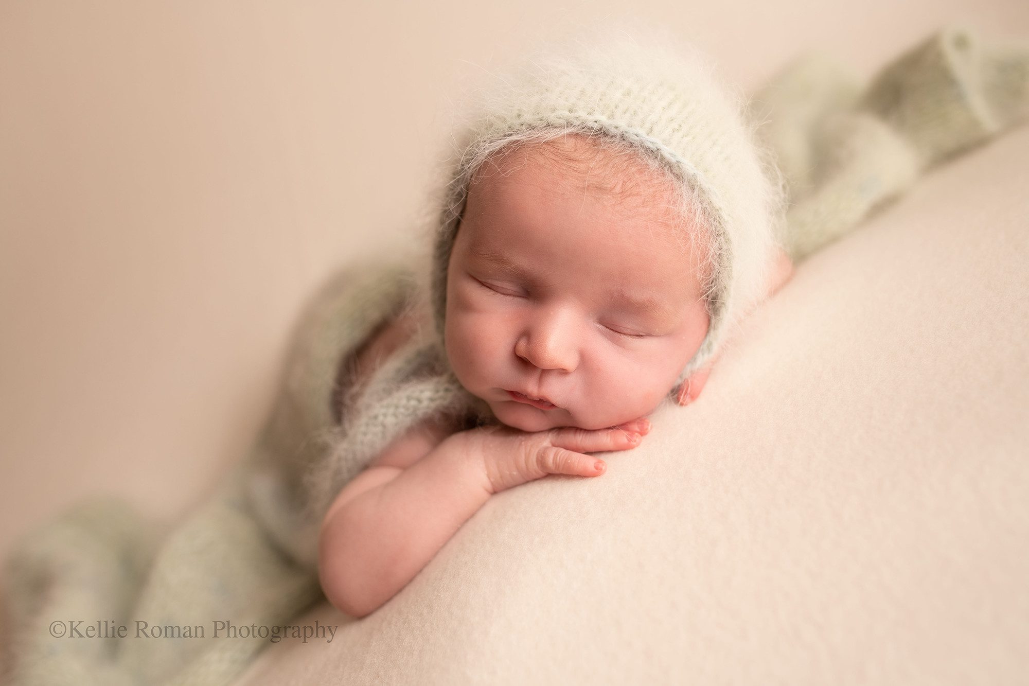 sweet and simple newborn. A newborn boy is in a milwaukee photo studio. He is laying on his belly on a cream backdrop with his hands under his chin. He is sleeping wand has a fuzzy light mint bonnet on, and a matching wrap covering his diaper.