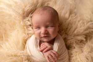 sweet and simple newborn. a baby boy is wrapped up in an ivory fabric, and is snuggled into ivory fur in a milwaukee photographers studio. his hand are sticking out of the wrap, and he is smiling.