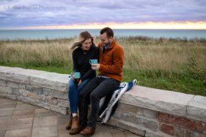 chilly sunrise a couple is at Lake Michigan during the sunrise. the couple is sitting on a brick wall with teal cups of coffee in their hands. the sky is cloudy with shades of purple, pink and blue. they couple is smiling and looking at each other