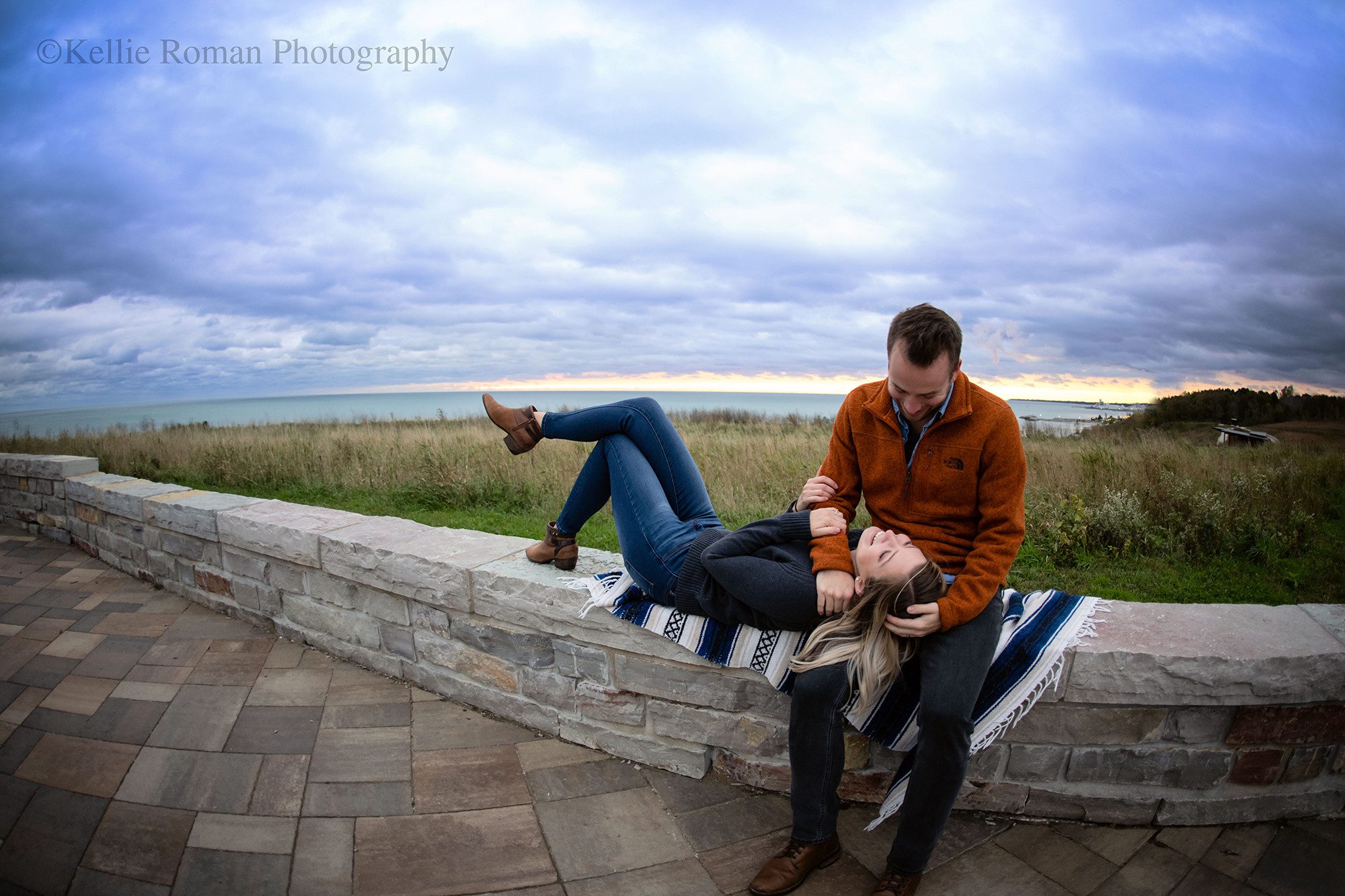 chilly sunrise a couple is at Lake Michigan during the sunrise. The man is sitting on a brick wall white his wife is laying with her head in his lap. the sky is cloudy with shades of purple, pink and blue. they couple is smiling and looking at each other