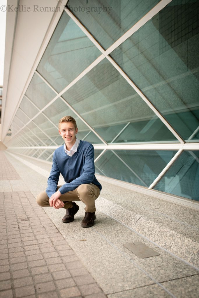 modern milwaukee senior. a milwaukee senior guy from greendale is in downtown milwaukee by the art museum. He's in front of a row of glass windows in front of the museum, and squatting down. He's wearing a blue sweater with khakis and is smiling at the camera.
