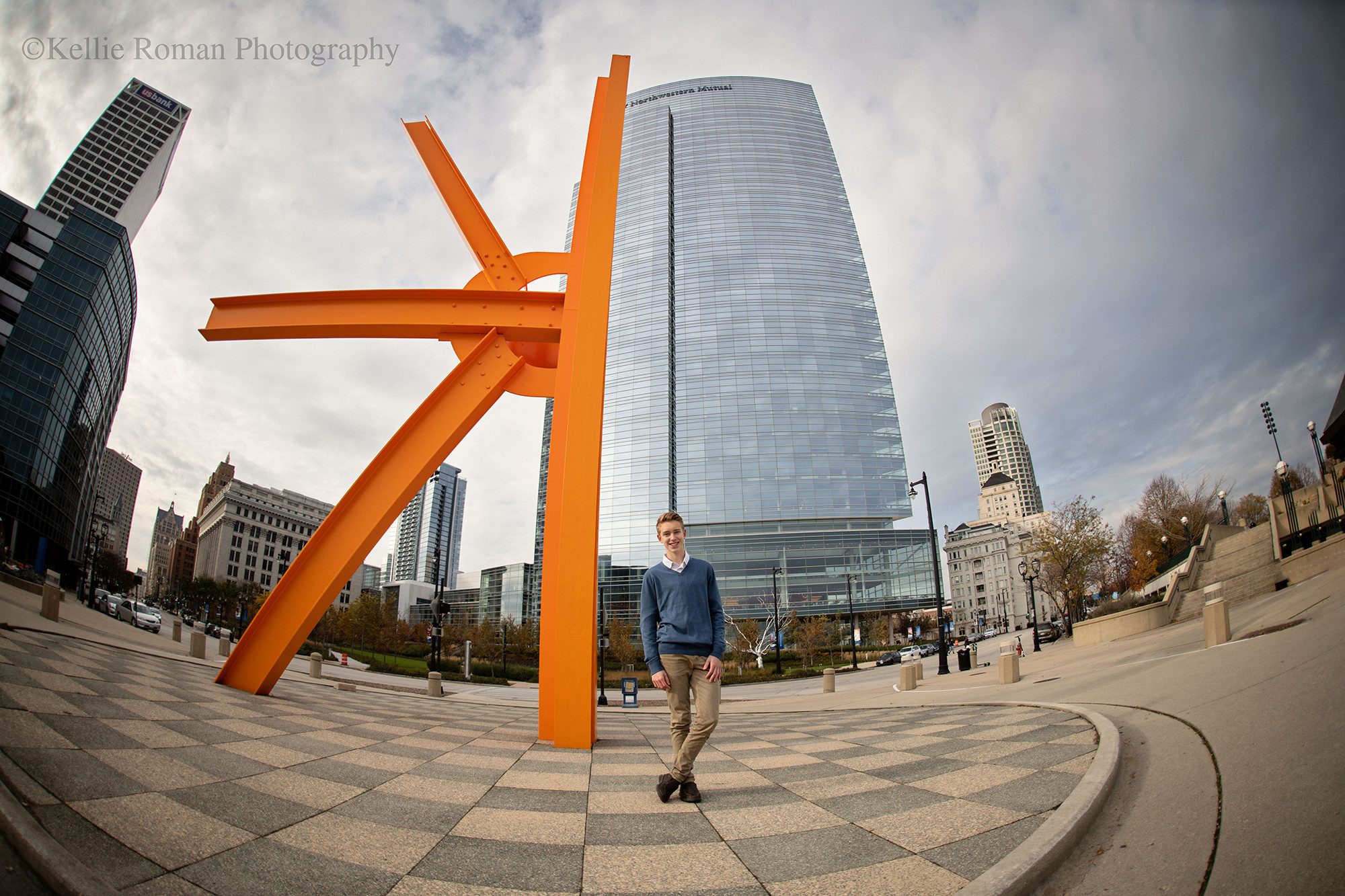 modern milwaukee senior. a high school senior boy from greendale is in downtown milwaukee. he's standing in front of an orange metal structure with the city of milwaukee behind him. the shot is very wide angle and gets the sky and all of the buildings within it.