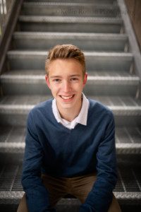 modern milwaukee senior. a greendale senior boy is in downtown milwaukee for his senior session. he's sitting on a metal staircase with his elbows on his knees smiling at the camera. he's wearing khakis and a blue sweater.