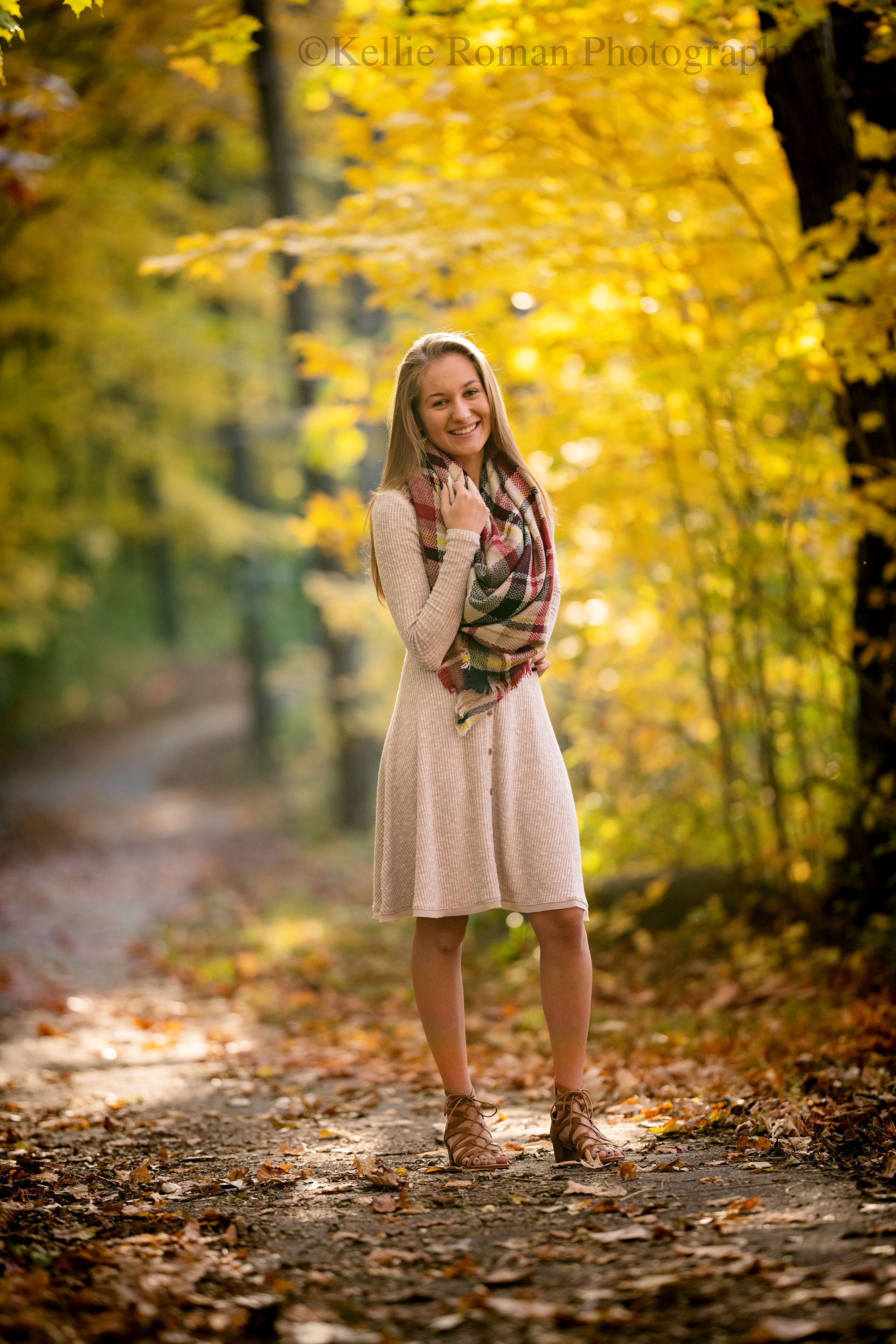 fall senior pics, a high school senior girl standing outside on a trail covered in yellow fall laves with yellow leaves and trees behind her. she's wearing a beige dress with a colored plaid scarf.