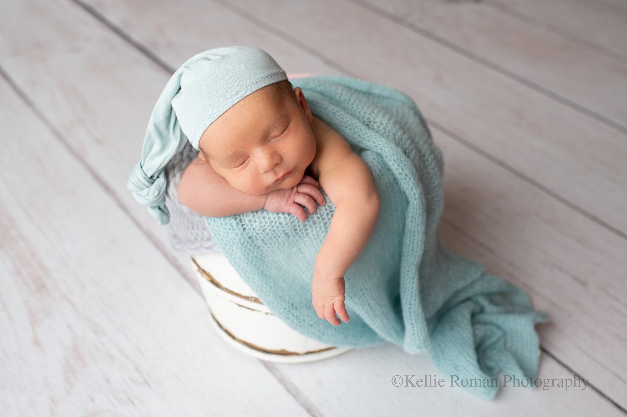 milwaukee photographer. newborn photography studio. Kellie Roman Photography. baby boy is sleeping in a white wood bucket that is stuffed with blue and grey fabrics. he has a light blue sleepy cap on. the newborn has one hand under his chin and the other hand is hanging over the edge of the bucket. 
