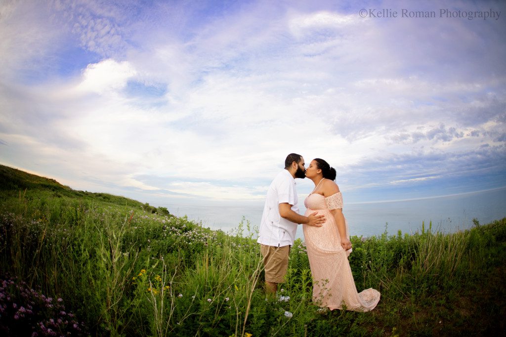 best greendale photographer. a women and her husband are standing outside during the summer on a grassy bluff overlooking Lake Michigan. there are yellow and purple wildflowers around them. they are leaning in and giving each other a kiss. the women is pregnant and is wearing a light pink lace dress. the man has a white polo on with khaki shorts. the sky is blue with pastel pink.