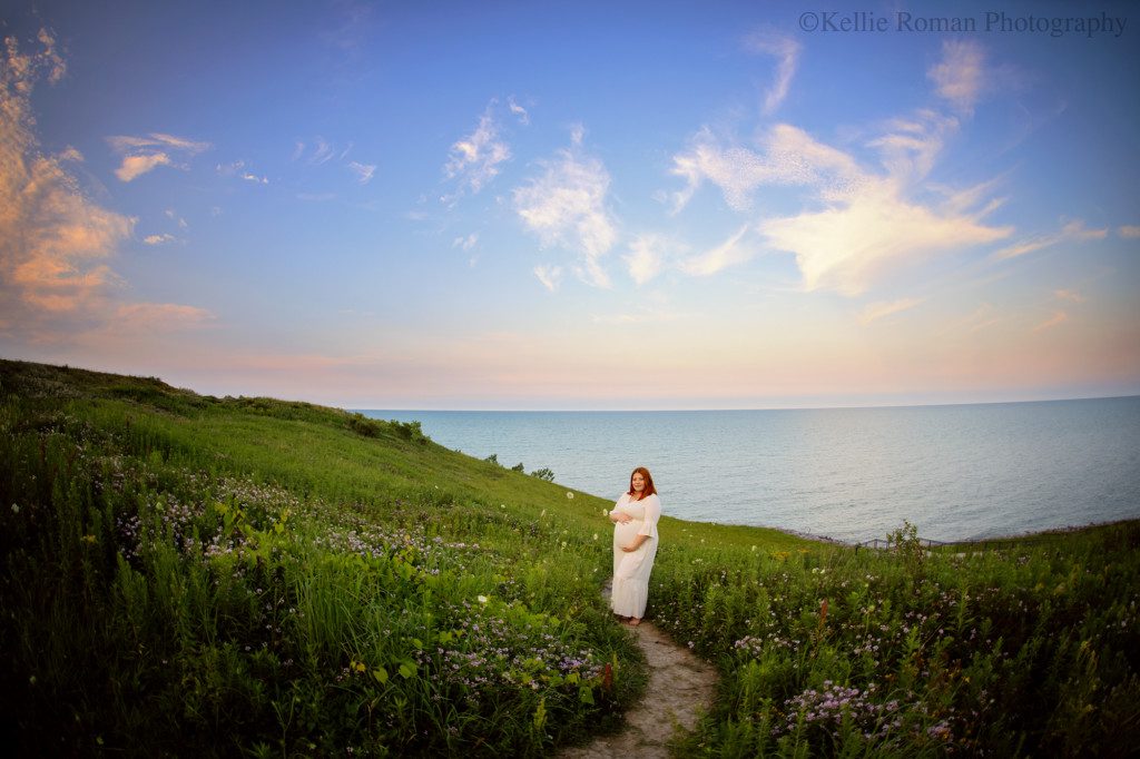 best maternity photographer. a pregnant women is standing on a bluff of tall green grass and wildflowers that overlooking blue waters of Lake Michigan in milwaukee. she is wearing a cream colored dress and holding onto her prengnant belly. the sky is pastel pinks and blues with some clouds.