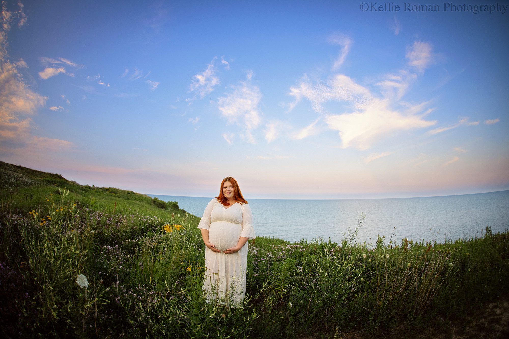 milwaukee maternity photographer. a pregnant soon to be mom is standing on a green grassy bluff overlooking Lake Michigan and a beautiful summer pastel sky. the women is smiling and looking into the camera. she is wearing a cream colored dress and is holding onto her belly. she's surrounded by wild flowers.