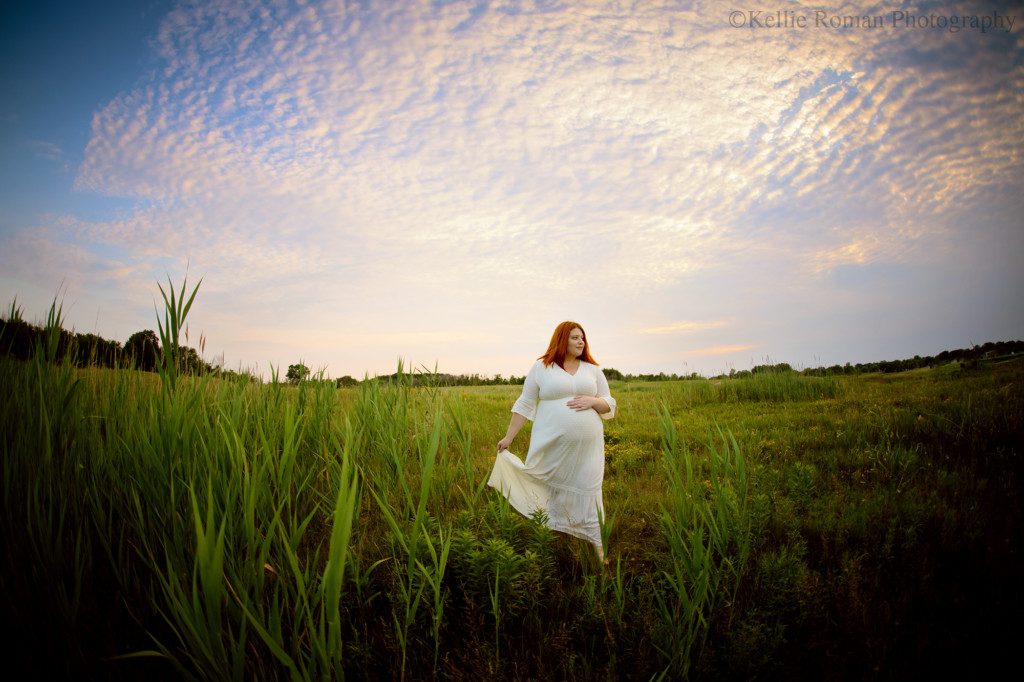 milwaukee best photographer. a pregnant women is walking through a field of tall grass with the peach sun setting behind her. she is wearing a cream flowing dress and has her hand holding up one side of her. her other hand is on her belly, and she's looking off to the side and smiling.