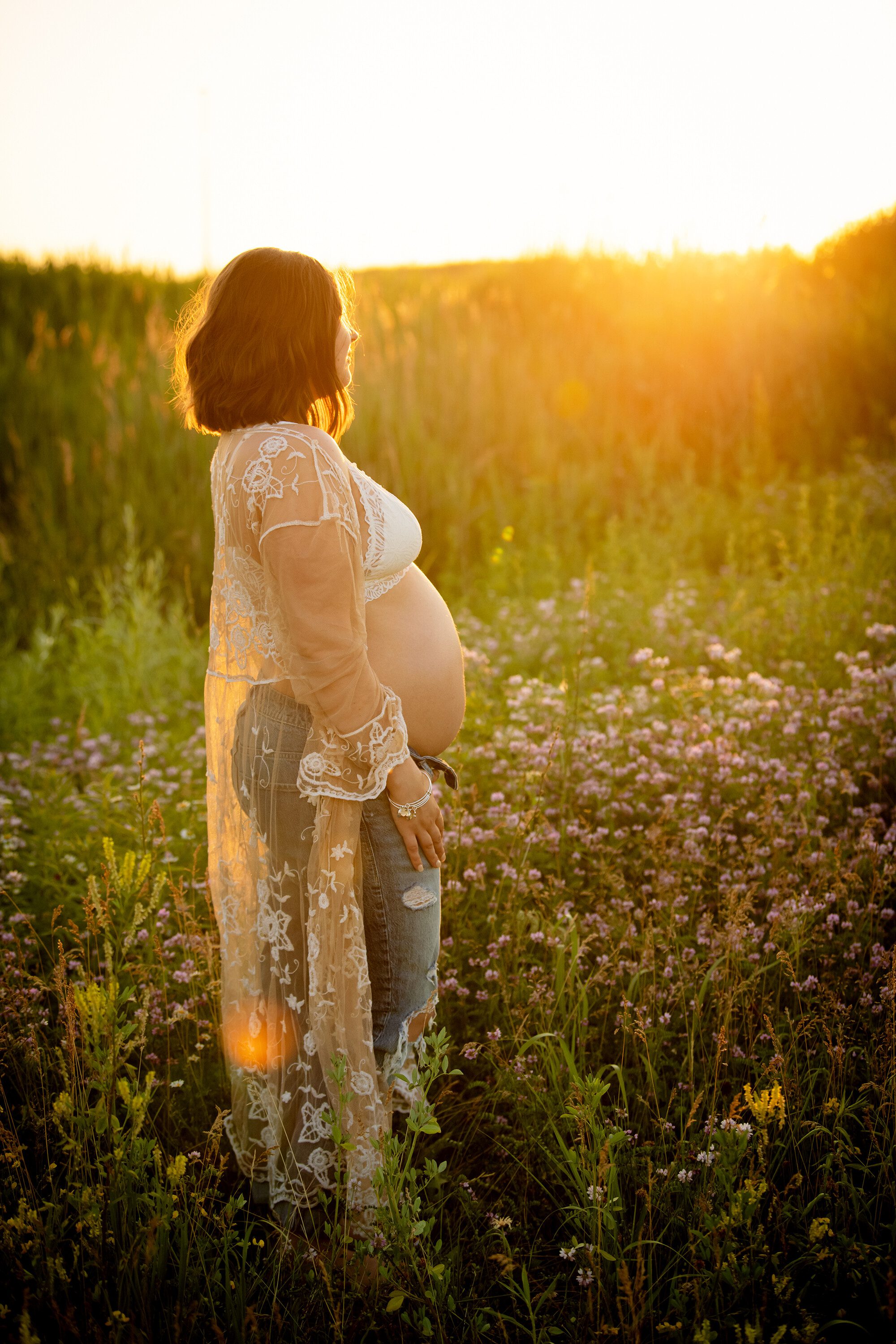 milwaukee maternity photographers. a pregnant women standing in a field of tall grass and wildflowers is looking in the distance with the golden sun setting behind her. she has on a white bra, with lace cover up exposing her pregnant belly.