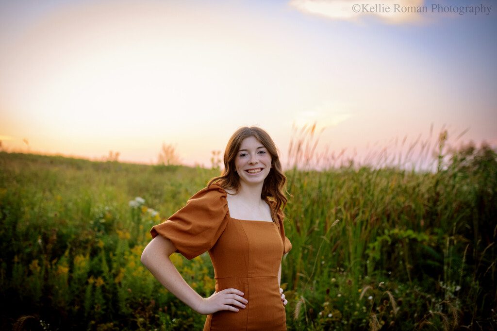 senior photography milwaukee. senior girl standing in milwaukee county park surrounded with green grass and wildflowers. the sun has set and sky is shades of pastel colors. girl has a burnt orange dress on and is standing with her hands on her hips.