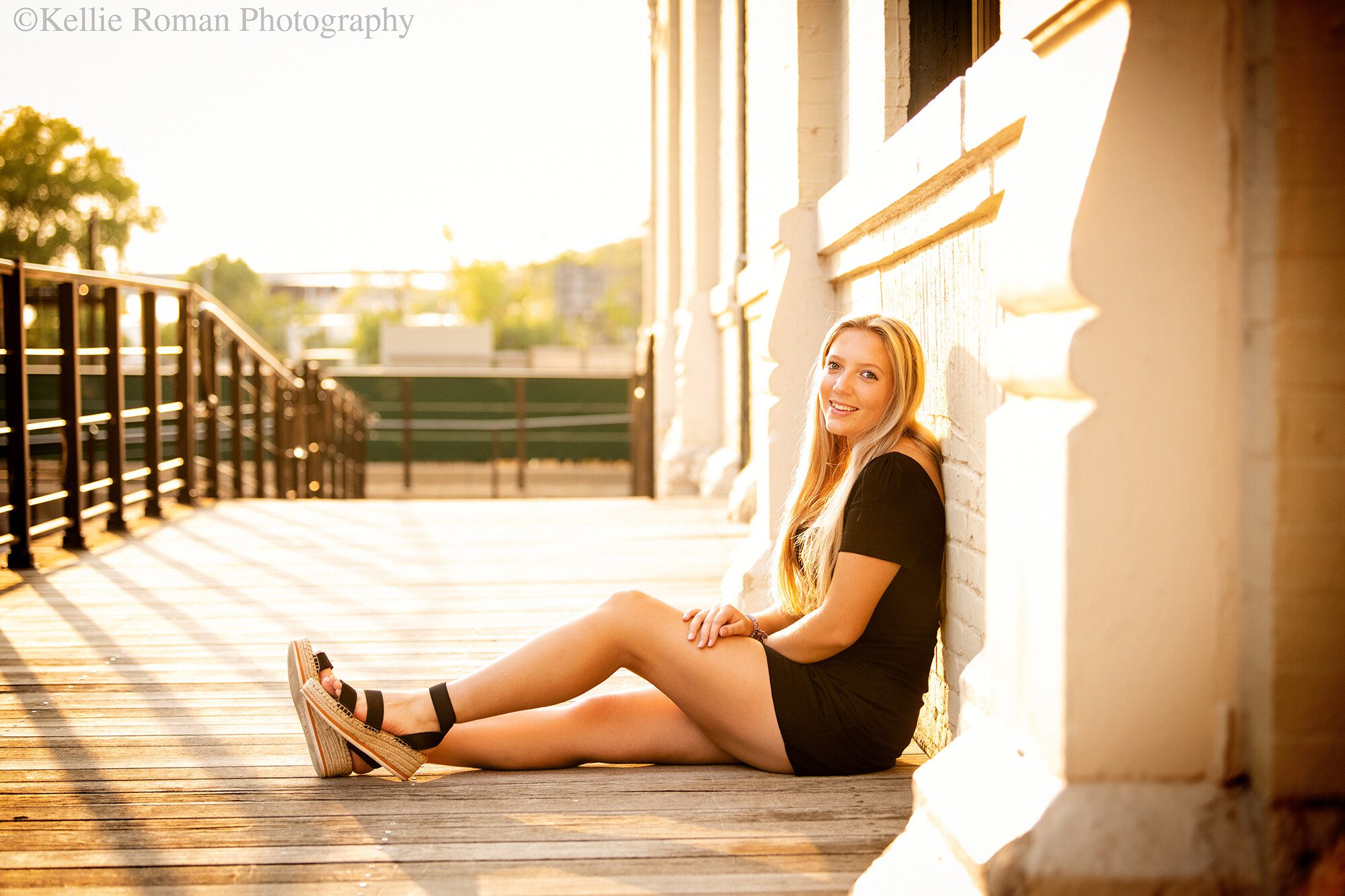 senior photos milwaukee. high school senior girl is sitting in downtown milwaukee with her back against a cream colored building. the ground is a wood boardwalk and the glowing golden summer sun is coming behind her. she has her legs out with hands resting in her lap. she's wearing a black dress and sandals.