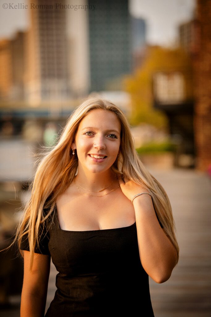 senior photographers milwaukee. a high school senior girl is in Milwaukee's downtown their ward on the river walk. the sun is setting to the side of her, and her long blonde hair is blowing in the wind. she's holding on side of her hair down with her hand while looking at the camera.
