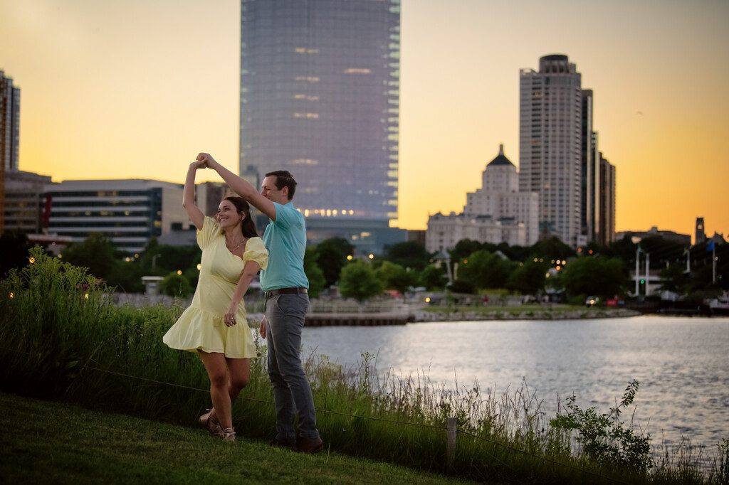 downtown milwaukee photographers. an engaged couple is in a park in front of the milwaukee skyline at dusk. the man is spinning the women around and she is wearing a light yellow dress. the man is in a blue polo shirt. they buildings have lights inside of them, and the sky is orange from the sun set.