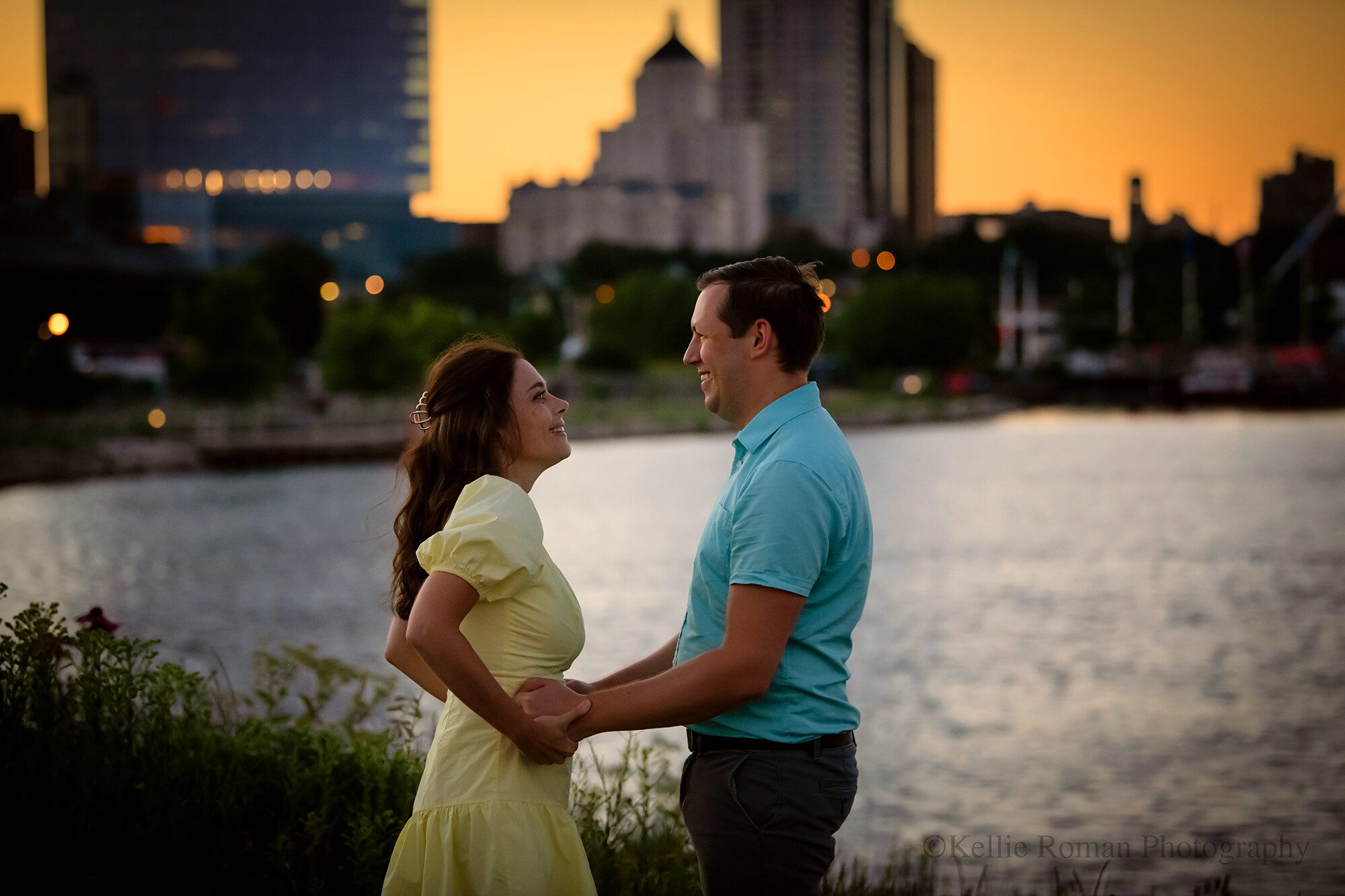 milwaukee photographer. Kellie Roman Photography. 
downtown milwaukee photography. an engaged couple is standing in a park in front of milwaukee skyline at dusk. the buildings are lit up and the sky is orange from the setting sun. they women is wearing a light yellow dress and is holding onto her fiancé's hands. the man is wearing a blue polo shirt and grey pants.