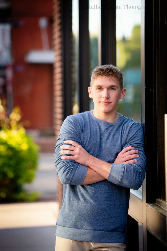 milwaukee senior portraits. milwaukee senior boy standing with arms crossed leaning against a black framed window in downtown milwaukee. boy has on a blue shirt and khaki pants and blonde hair.