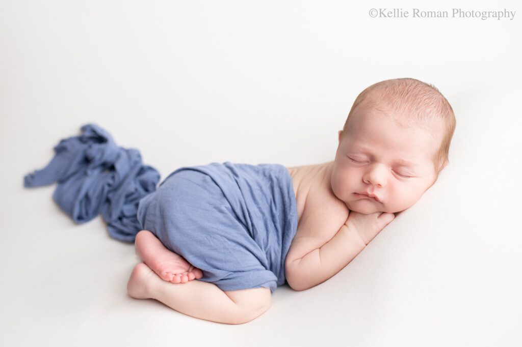 pastel newborn photographer. a newborn baby boy is in greendale photography studio. he is sleeping on his belly on top of a cream fabric. he's curled up with his chin resting on his hands and his feet showing. he has a blue swaddle wrap cover his body.