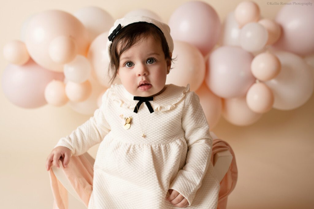 one year old girl in studio cake smash. girl has on a cream dress with black neck tie and necklace. she's wearing a matching beret and has big green eyes with dark hair. she's sitting on a white bench and staring at the camera. the backdrop is a balloon garland with blush pink and cream balloons. 
