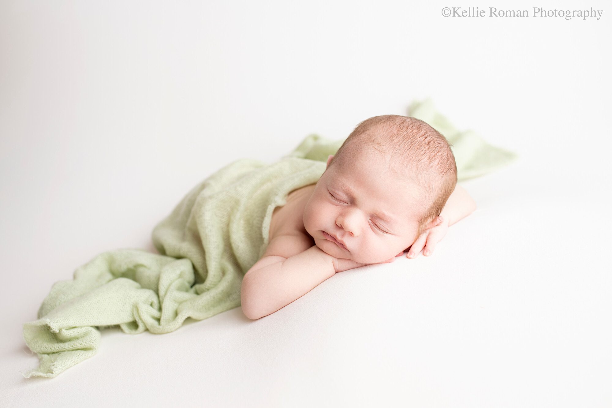 milwaukee newborn pics. a baby boy is in greendale photography studio. he is sleeping on his belly with his chin resting on his hands. he's laying on top of a cream fabric, with a light green knit swaddle covering his diaper, feet, and back.