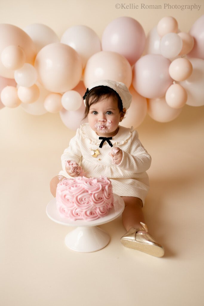 one year old girl in milwaukee studio cake smash session. girl is wearing a cream dress and beret hat with gold shoes. she's infront of a light pink cake in a white cake stand. the backdrop is a balloon garland with cream and blush pink balloons.