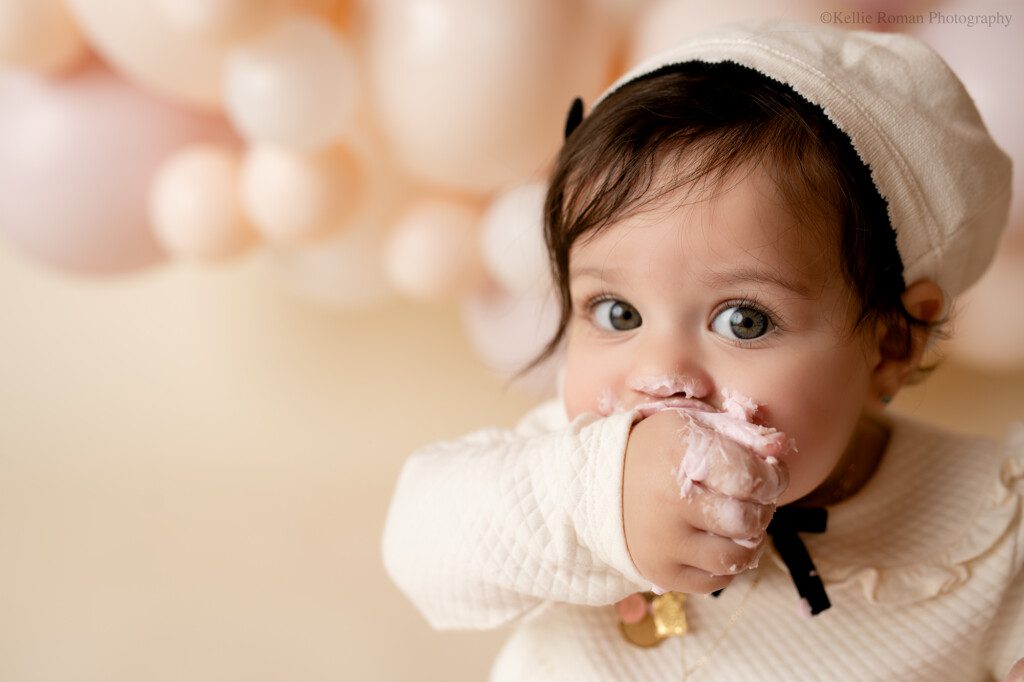 one year old girl in milwaukee studio cake smash session. girl has dark hair and a cream beret and outfit on. the backdrop are balloons of neutral cream and blush pink, she has frosting on her hand and her hand is in her mouth.
