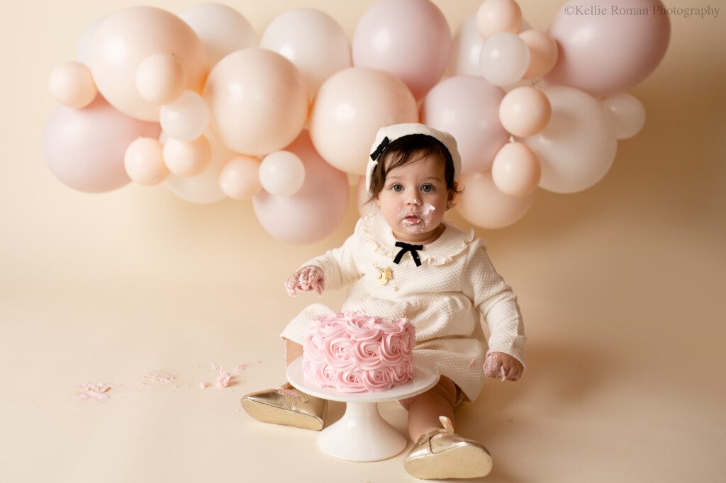 one year old girl in milwaukee studio cake smash session. girl is wearing a cream dress and beret hat with gold shoes. she's infront of a light pink cake in a white cake stand. the backdrop is a balloon garland with cream and blush pink balloons. she has pink frosting on her face and her hands. 