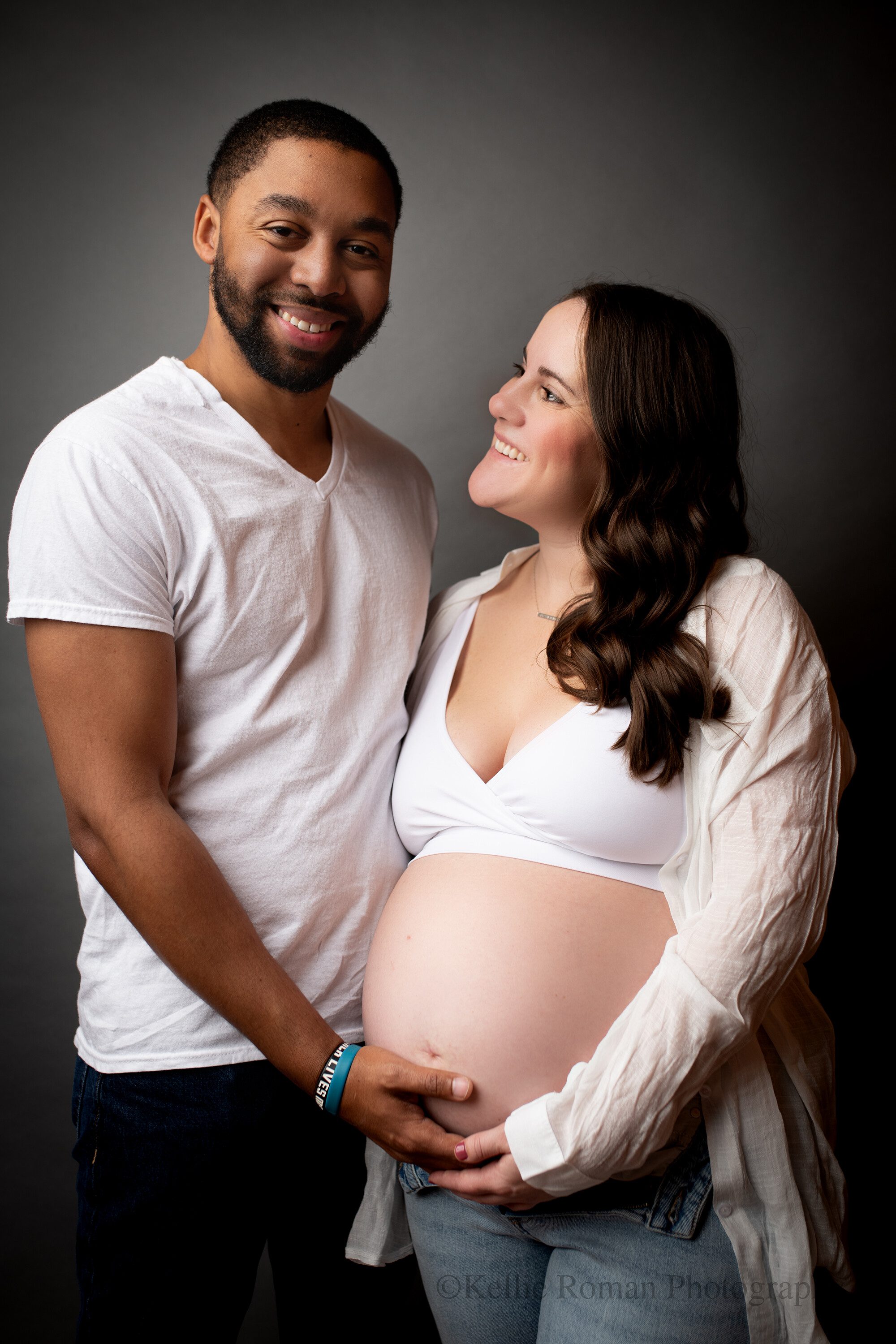 milwaukee area photographersa husband and wife in milwaukee photographer studio. they are wearing white t shirts and jeans, smiling and looking at the camera. the women is pregnant with child, and both husband and wife have their hands on her exposed baby belly.