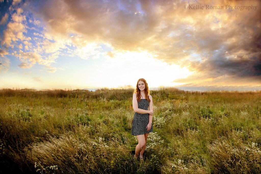 milwaukee seniors. senior girl standing outside in field of tall grass with beautiful sun set behind her. sky has purple and blue clouds and girl has red curly hair with black and white dress on.
