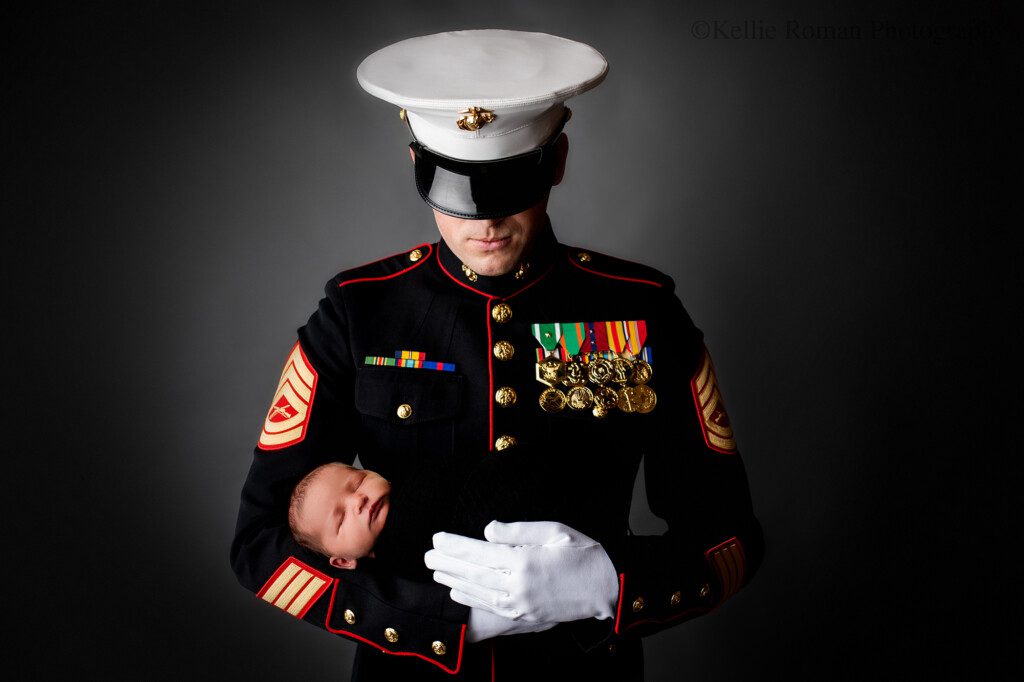 best milwaukee photographers. milwaukee newborn photography studio has a man dress in his United States Marine dress uniform holding his newborn baby who is wrapped in a black swaddle. the marine father is looking down, and is standing in front of a dark grey backdrop.