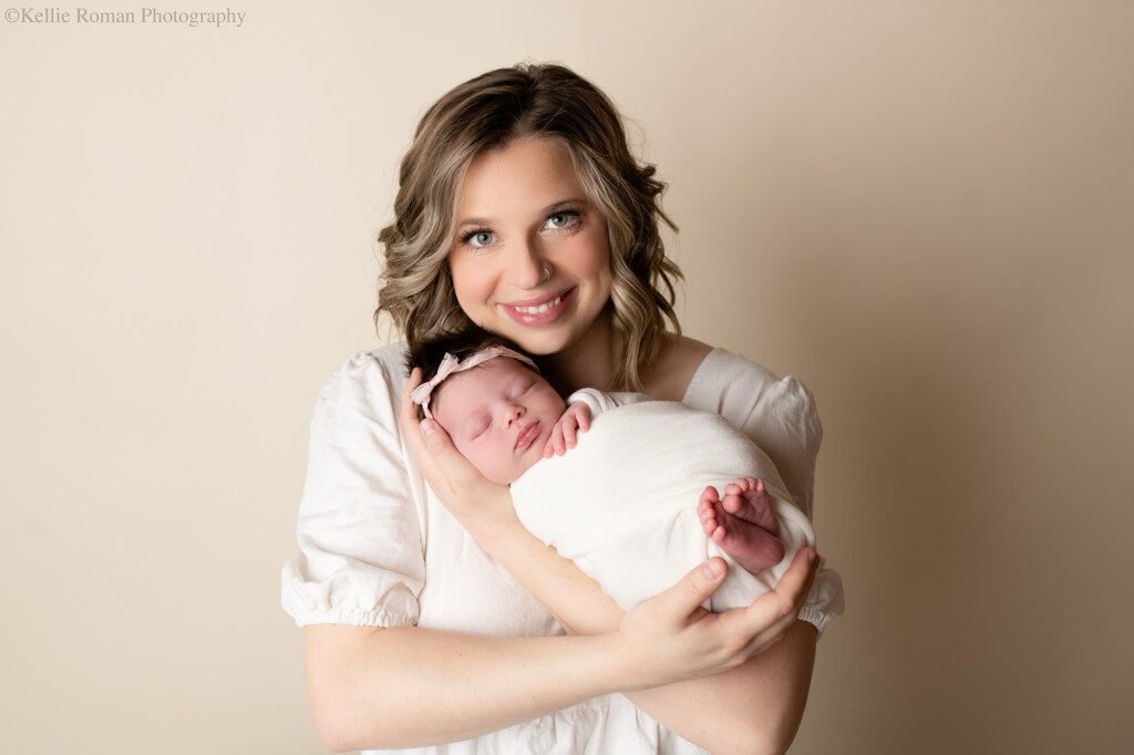 milwaukee newborn pics. a new mother is holding her newborn baby girl while smiling at the camera. the newborn baby is wrapped in a cream swaddle with her feet sticking out. the backdrop is cream colored, and the mom has a cream dress on.