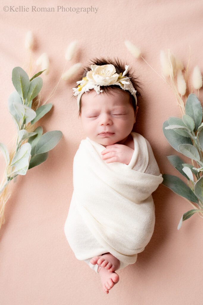 best newborn photographers. milwaukee newborn in greendale photo studio. baby girl is wrapped in a cream fabric laying on top of pink fabric. she is surrounded by flowers and has a floral headband on.