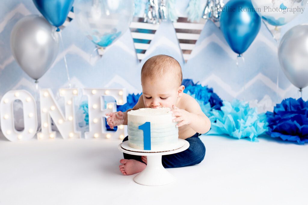 cake smash milwaukee. a one year old boy is in milwaukee photo studio having his first cake smash for his birthday. the backdrop is a chevron light blue and white with silver and blue banner, and silver and blue balloons. tissue paper pom moms are on the floor in different shades of blue, and a white marquee letters that spell out one. the boy has no shirt on and jeans, and has his face biting into his blue and white smash cake.