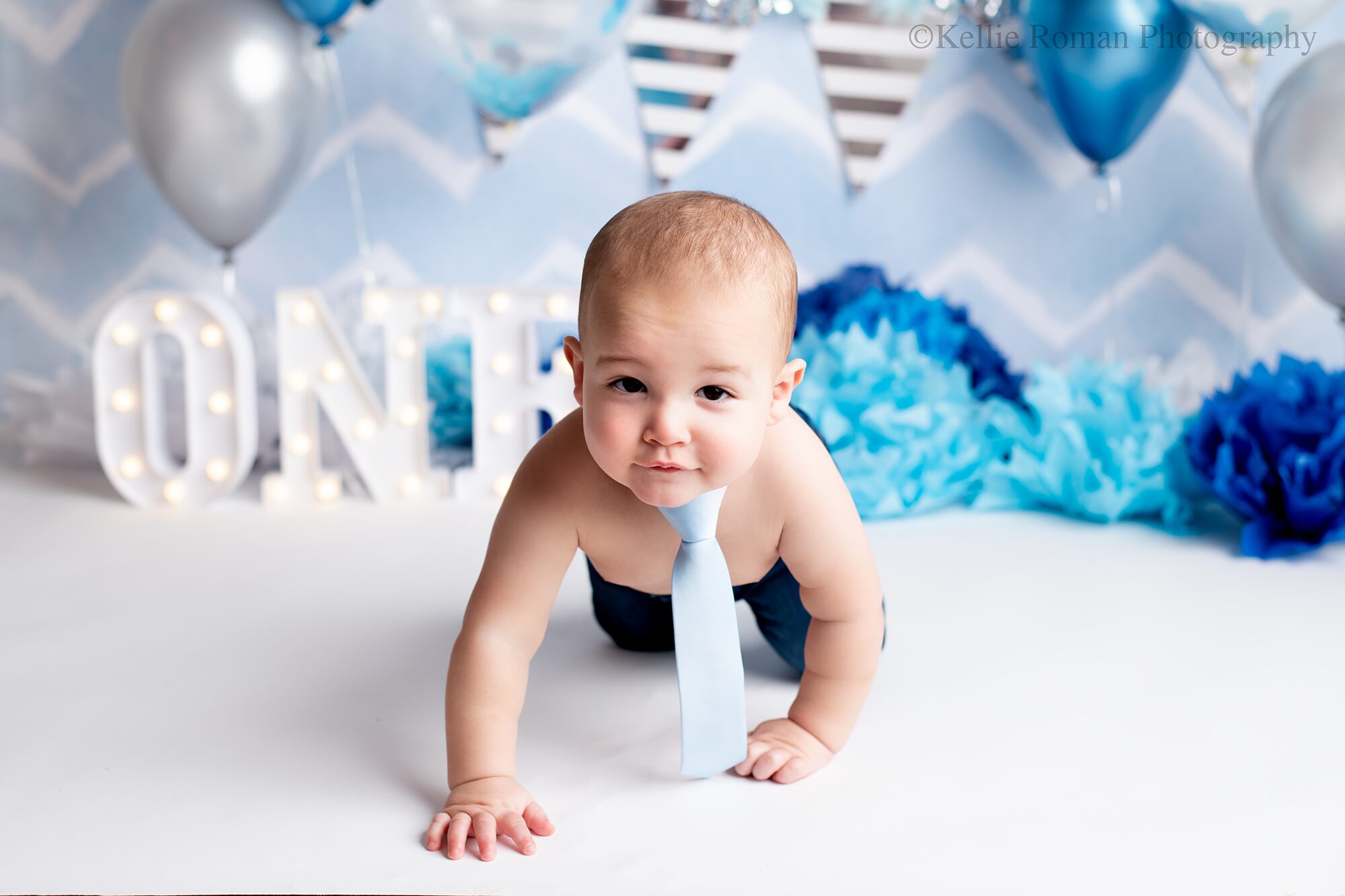 greendale cake smash. one year old boy in photo studio. he's crawling and looking at the camera. he has a baby blue tie and jeans on with no shirt. the backdrop is baby blue white and silver.