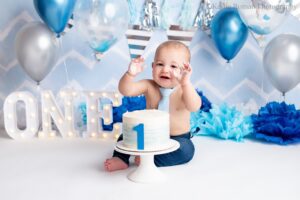 greendale cake smash. one year old boy in photo studio is sitting behind a blue and white smash cake on a white cake stand, the cake has a blue number one on the front. boy has jeans and a baby blue tie on with no shirt. the backdrop is blue and white chevron with silver and blue balloons and banners.