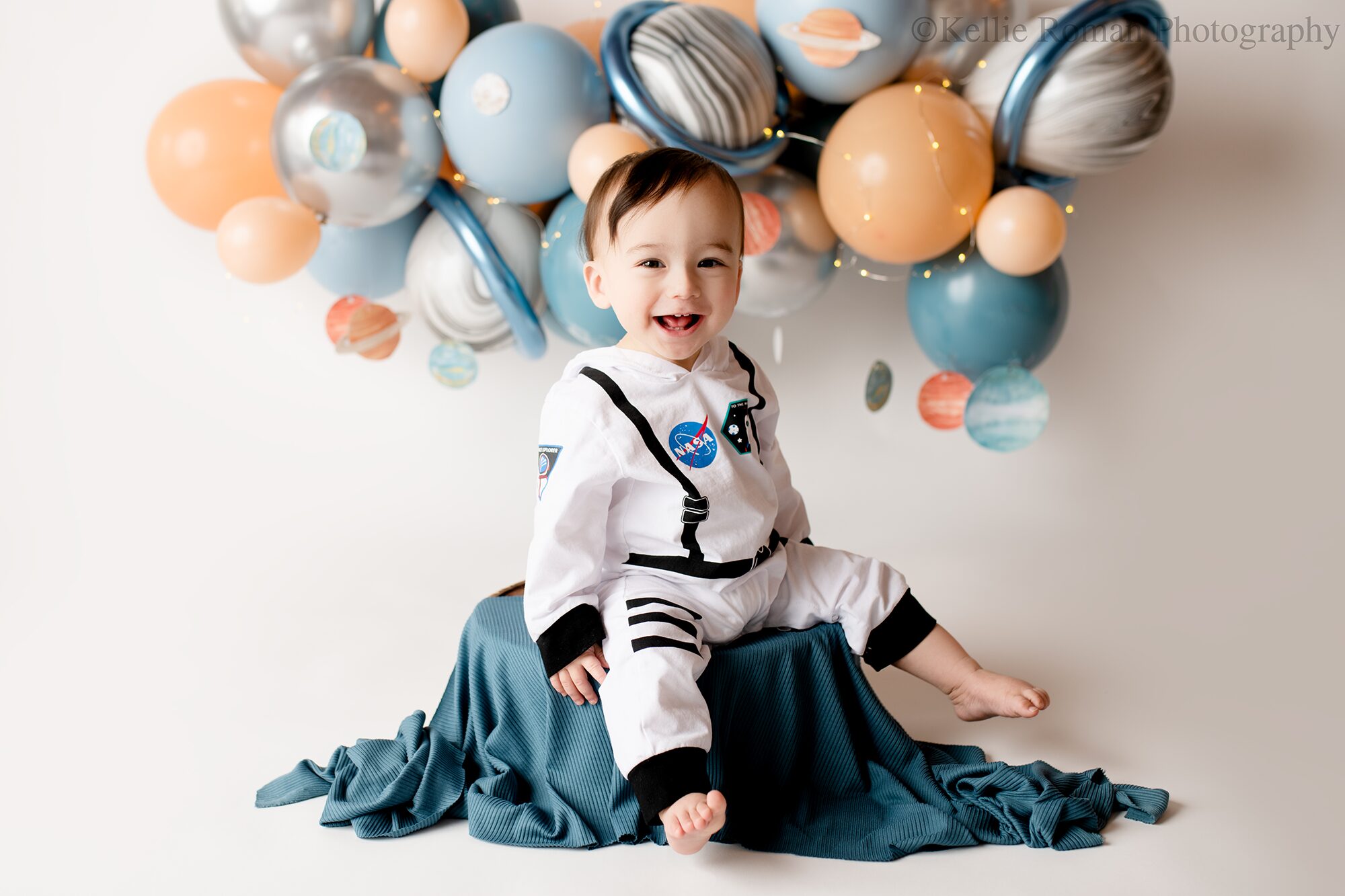 space cake smash. one year old boy in greendale photography studio. the backdrop is white with a blue, silver, and beige balloons garland. there are small lights and planets on the balloons. the boy is sitting on a wood tub, with a blue fabric over it. he's wearing a space onsie and smiling.